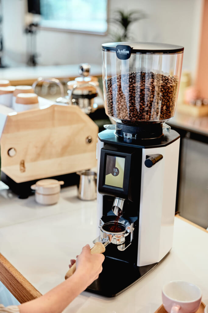 White espresso grinder with a glass bean hopper full of roasted beans a hand holds a portafilter underneath to collect grounds