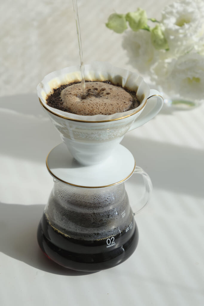 Pour over coffee brewing hot water streams over coffee grounds in a white porcelain dripper with gold trim above a glass carafe