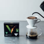 Pour over coffee setup with a white ceramic dripper on a carafe hot water pouring from a black kettle a Hario V60 filter box nearby