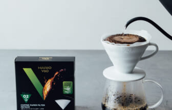 Pour-over coffee setup with a white ceramic dripper on a carafe, hot water pouring from a black kettle, a Hario V60 filter box nearby.