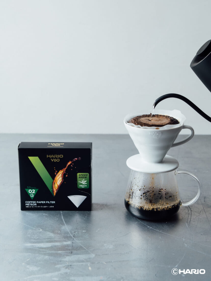 Pour over coffee setup with a white ceramic dripper on a carafe hot water pouring from a black kettle a Hario V60 filter box nearby