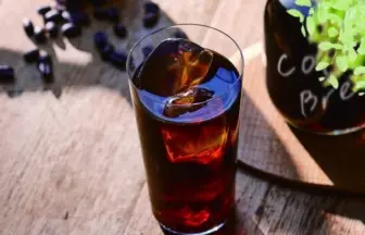 Tall glass of iced dark cola on a sunlit wooden table, with ice cubes visible and reflections on the surface.