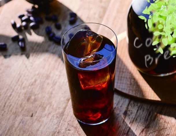 Tall glass of iced dark cola on a sunlit wooden table with ice cubes visible and reflections on the surface
