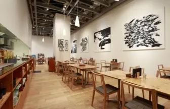 Bright modern cafe interior with wooden tables and chairs, pendant lights, and large black-and-white wall art on white walls.