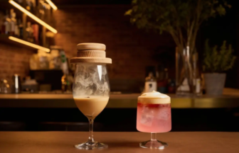 Smoky cocktail in a tall stem glass with a wooden lid beside a pink foamy cocktail on a bar counter