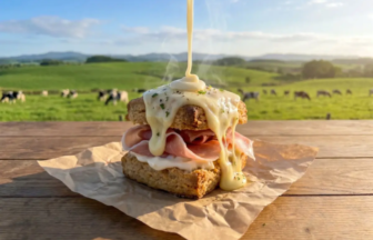 Sandwich with melted cheese and ham on rustic bread, cheese sauce dripping, outdoors on a wooden table.