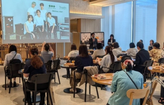 Event in a bright room with a large screen displaying a group photo two presenters speak at a lectern to an audience seated at round tables