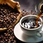 Freshly brewed coffee pours into a white cup from a glass pot with a burlap sack of coffee beans nearby