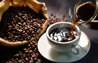 Freshly brewed coffee pours into a white cup from a glass pot with a burlap sack of coffee beans nearby