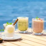 Three glass mugs with cold drinks on a sunlit wooden table by the sea lemon mint drink green smoothie and pink berry smoothie with a small syrup jug nearby