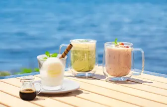 Three glass mugs with cold drinks on a sunlit wooden table by the sea lemon mint drink green smoothie and pink berry smoothie with a small syrup jug nearby