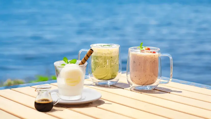 Three glass mugs with cold drinks on a sunlit wooden table by the sea lemon mint drink green smoothie and pink berry smoothie with a small syrup jug nearby