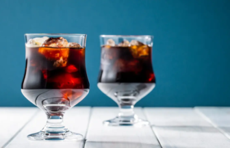 Two stemmed glasses of dark cola with ice on a white wooden table against a teal background