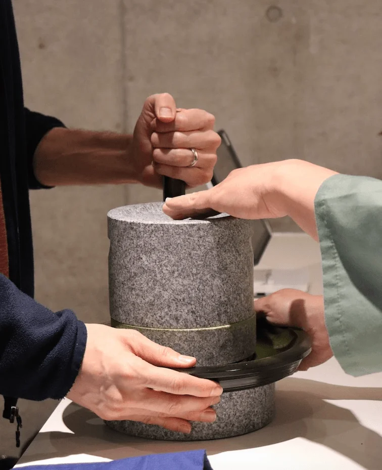 石臼を使って抹茶などの材料を挽いている様子を捉えた画像です。二人の手が協力し、伝統的な方法で丁寧に作業を進めている光景が伺えます。