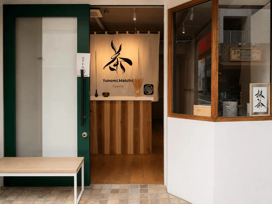 抹茶専門店「Yunomi.Matcha TOKYO」の店舗入口です。緑色の扉と「茶」の文字が入った暖簾が特徴的で、和モダンな雰囲気が漂います。店内には木製カウンターが見え、外にはベンチが置かれています。