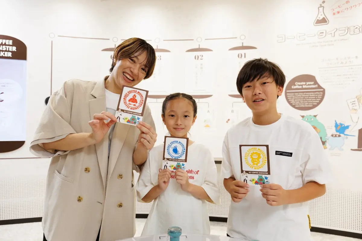 UCCコーヒーミュージアムで、大人と子供2人が「マイコーヒーモンスター」のカードを持って笑顔で写っている。コーヒーの未来を体験できるイベントの様子。