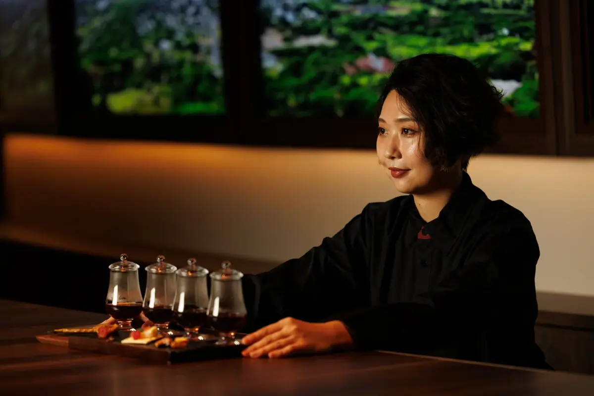 黒いシャツを着た女性が、木製のトレーに載せられた4つのグラスに入った飲み物と軽食のテイスティングセットを前に座っています。背景には緑豊かな景色が見え、落ち着いた雰囲気です。
