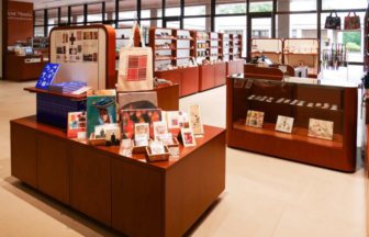 Interior of a bookstore with wooden display tables, shelves of books and postcards, and a tote bag hanging from a display post in the background.