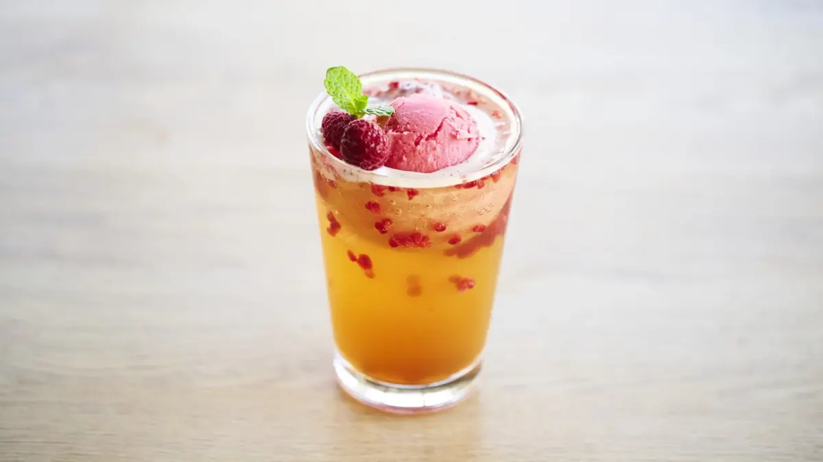 明るい木目のテーブルに置かれたグラスに入った冷たい飲み物。オレンジ色のドリンクの上にピンクのアイスクリームが乗っており、フレッシュなラズベリーとミントの葉が添えられています。飲み物の中にもラズベリーの粒が見えます。