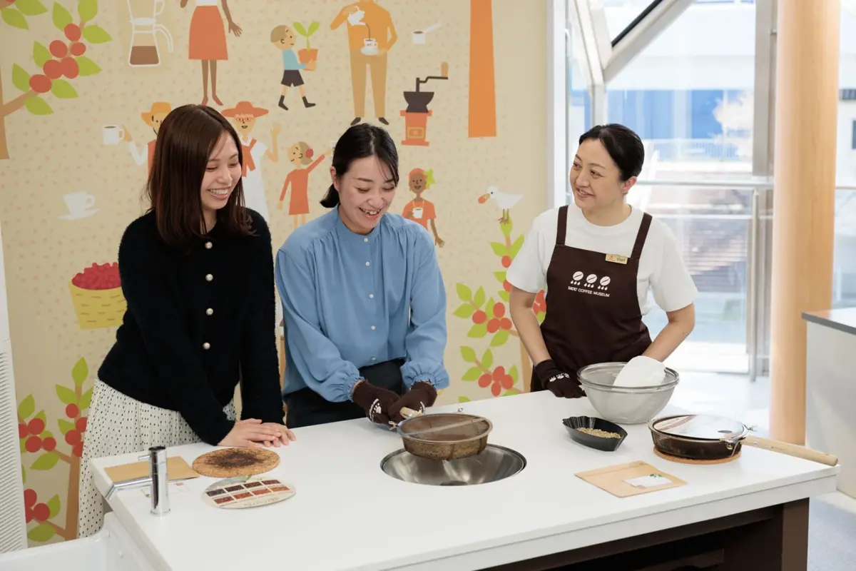 コーヒー博物館で3人の女性がコーヒー豆の焙煎ワークショップに参加している様子。笑顔で楽しそうに作業しており、テーブルにはコーヒー豆や焙煎器具が並べられている。背景にはコーヒーをテーマにしたカラフルな壁画がある。