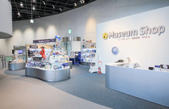 Museum Shop interior with curved white counters, circular display island, and shelves of souvenirs under bright lighting, staff in red nearby.