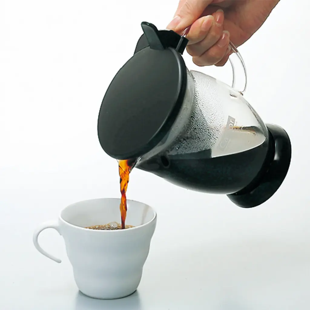 Person pours hot coffee from a glass kettle into a white mug on a white surface