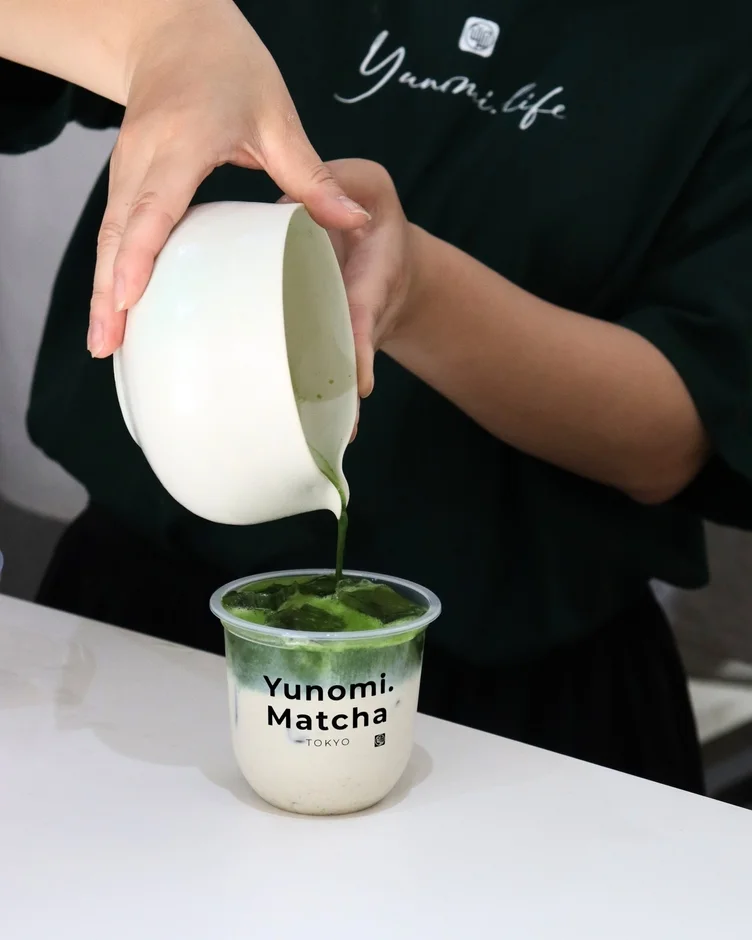 白いピッチャーから鮮やかな緑色の抹茶が、牛乳と氷が入った透明なカップに注がれている様子。カップには「Yunomi. Matcha -TOKYO」と書かれており、抹茶ラテが作られている瞬間を捉えています。