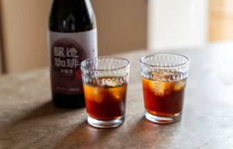 Two short glasses with dark iced beverages and ice cubes on a wooden table, a bottle with Japanese characters in the background.