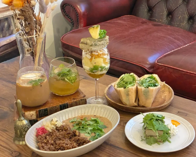 Table spread of drinks and dishes two creamy beverages a layered fruit yogurt parfait two veggie wraps a bowl of lentil and rice and a small herb topped bite on a plate with a leather sofa in the background