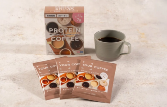 Box labeled PROTEIN COFFEE with sachets and a gray mug of coffee on a light surface four brown packets stand in front of the box