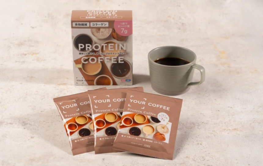 Box labeled PROTEIN COFFEE with sachets and a gray mug of coffee on a light surface four brown packets stand in front of the box
