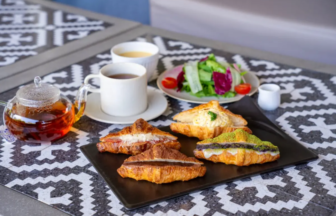 Croissants on a black platter with a teapot, two cups of tea, and a side salad on a patterned outdoor table.