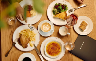 Top down view of a brunch table with plates of fries and roasted chicken a salad a slice of quiche crème brûlée coffee water and wine and a black menu on wood