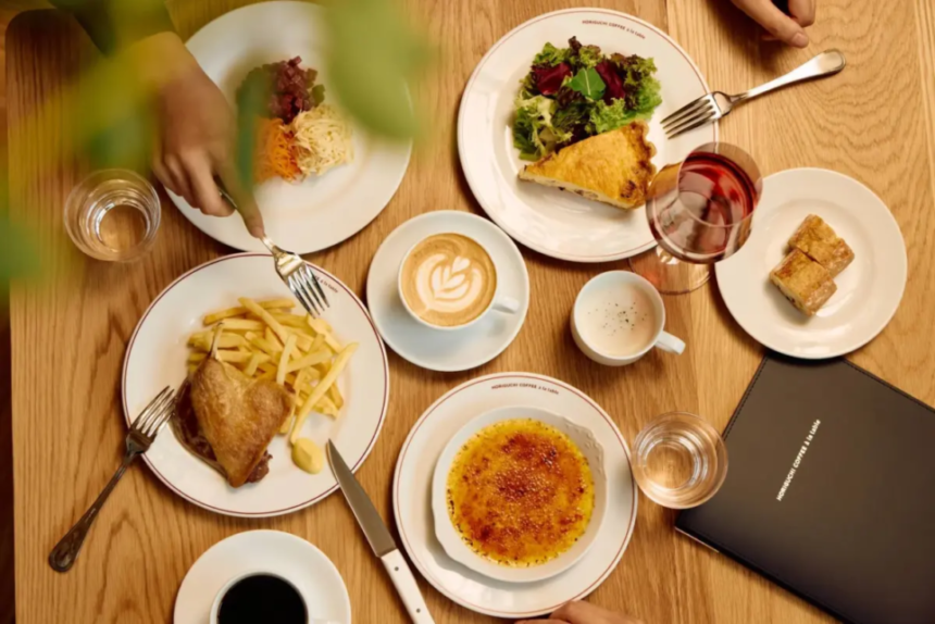 Top down view of a brunch table with plates of fries and roasted chicken a salad a slice of quiche crème brûlée coffee water and wine and a black menu on wood