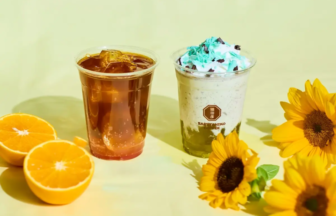 Two clear cups with drinks (iced coffee and a dessert with blue topping) sit on a yellow table beside orange halves and bright sunflowers.