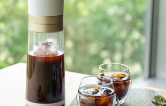 Iced coffee dispenser with white lid and beige band beside two glasses of iced coffee on a wooden table with cookies nearby and greenery outside the window