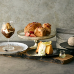 Assorted desserts on a rustic board chocolate parfait in a glass berry topped buns a lemon cake slice and a small glass of liqueur