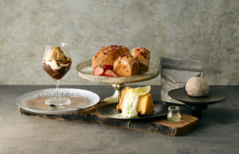 Assorted desserts on a rustic board: chocolate parfait in a glass, berry-topped buns, a lemon cake slice, and a small glass of liqueur.