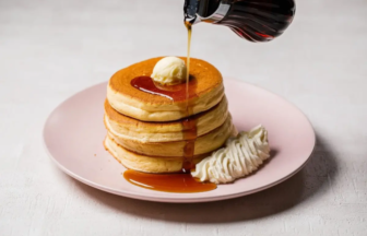 Stack of fluffy pancakes with a pat of butter, maple syrup being poured over them on a pink plate.