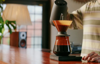Person making pour-over coffee at a wooden kitchen counter, pouring hot water through a cone filter into a glass carafe.