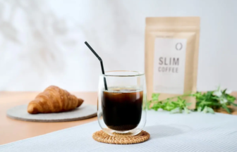 Glass of iced coffee with a black straw on a woven coaster croissant on a plate and a Slim Coffee bag in the background