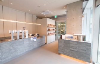 Bright, modern cafe or shop interior with gray stone counters, coffee equipment, and product shelves along the wall near a concrete pillar.