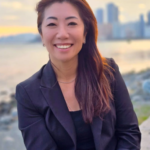 Smiling woman in a black blazer posing outdoors by a waterfront at sunset with a city skyline in the background