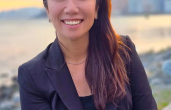 Smiling woman in a black blazer posing outdoors by a waterfront at sunset with a city skyline in the background