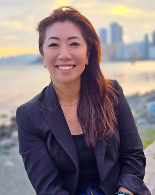 Smiling woman in a black blazer posing outdoors by a waterfront at sunset with a city skyline in the background