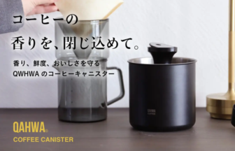 Black QAHWA coffee canister on a wooden surface with a hand placing a filter into a grinder in the background, text promotes keeping coffee aroma and freshness.