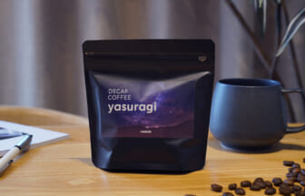 Bag of decaf Yasuragi coffee on a wooden table beside a gray mug with scattered coffee beans nearby