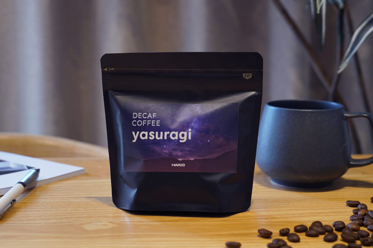 imageyasuragi | コーヒーステーション Bag of decaf Yasuragi coffee on a wooden table beside a gray mug with scattered coffee beans nearby