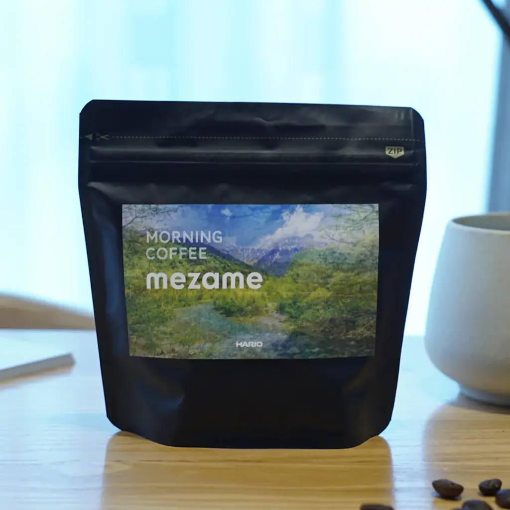 mezameimagesquare | コーヒーステーション Black coffee bag on a table labeled Morning Coffee mezame with a scenic label coffee beans nearby and a cup in the background