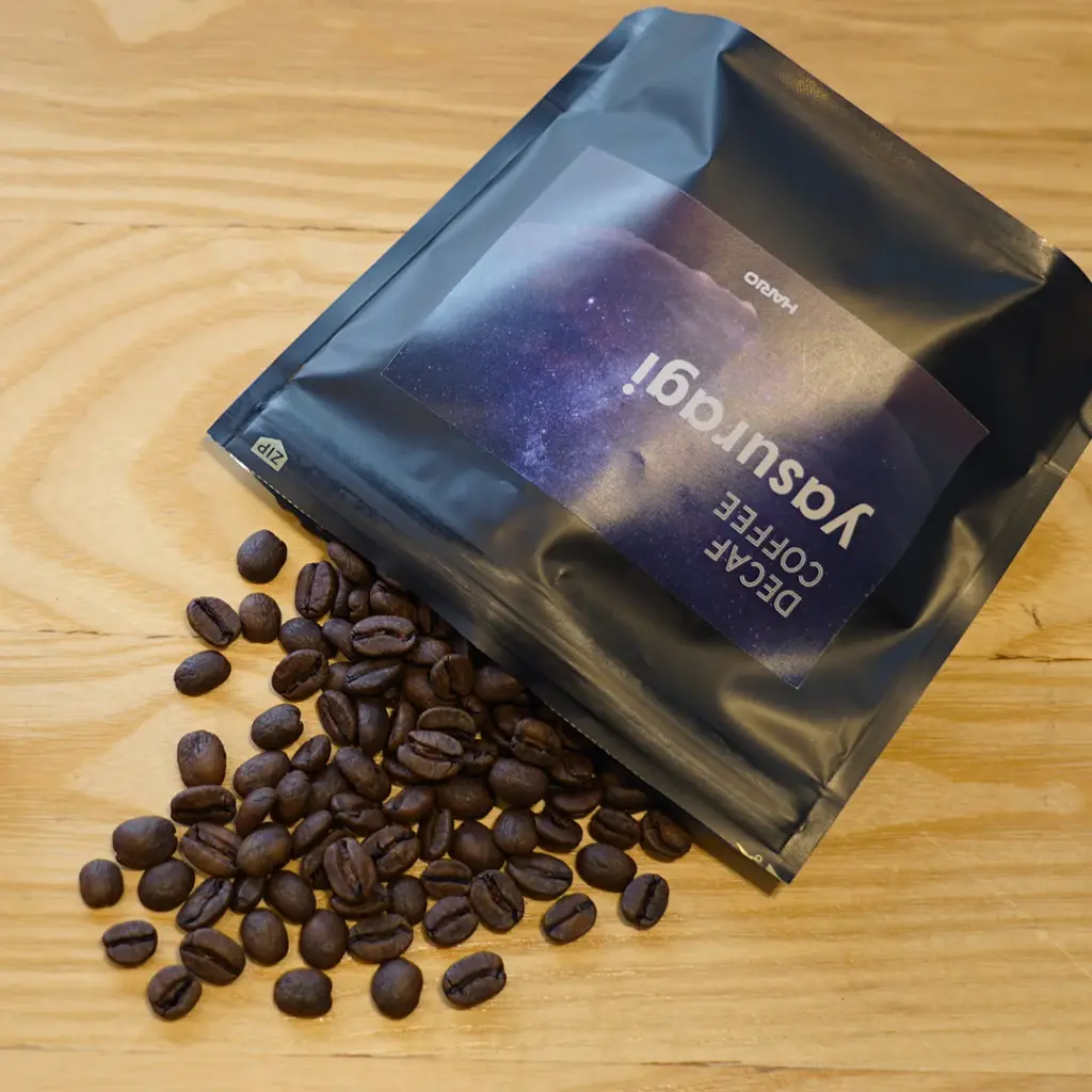 yasuragimame2 | コーヒーステーション Bag of roasted coffee beans spilled onto a wooden surface beside a dark resealable coffee bag